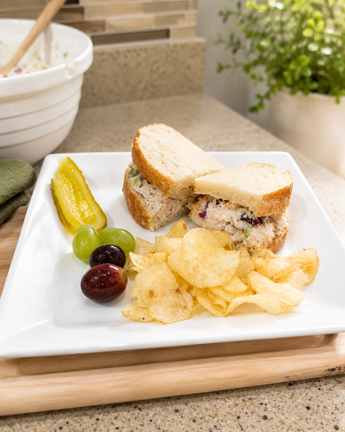 Cranberry walnut chicken salad sandwich on soft bread served with a dill pickle spear, grapes, and kettle chips on a white plate